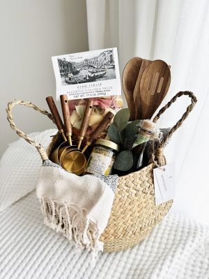 Luxury gourmet gift basket titled The Artisanal Kitchen by Divine Bloom Sarasota, featuring gold and wood measuring cups, authentic pasta, premium basil pesto, and eucalyptus accents in a woven wicker basket.