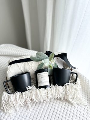 Luxury monochromatic modern gift box titled The Noir by Divine Bloom Sarasota, featuring two matte black ceramic mugs, a Jasmine Santal soy candle, and a cream waffle knit throw blanket with a black satin bow.